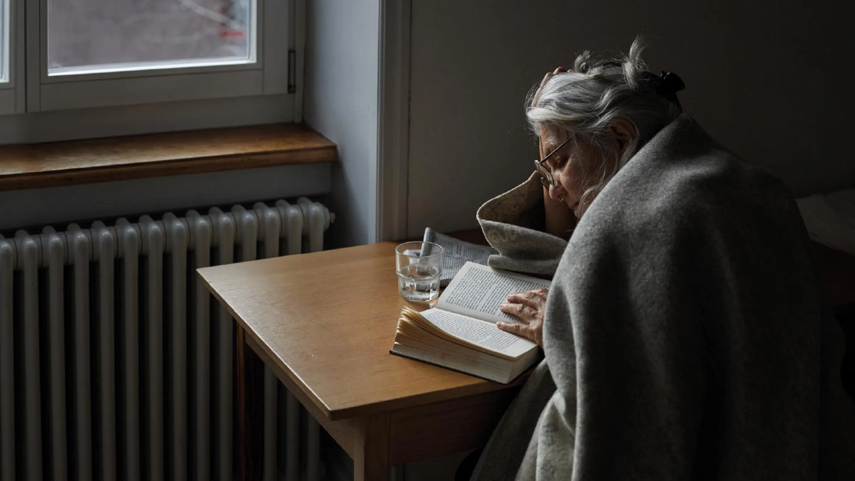 Une femme âgée est assise à une table et lit un livre.